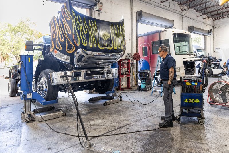 Technician inspecting lifted heavy-duty diesel truck with hood open inside repair shop, performing maintenance using diagnostic equipment and tools advanced